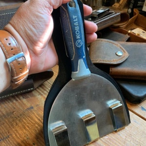 May include: A black leather tool holder with three metal clips. A blue and grey putty knife with the word "KOBALT" printed on the handle is inserted into the holder.