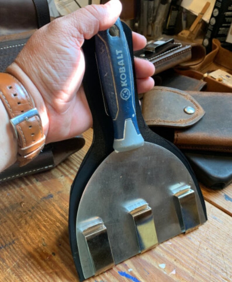 May include: A black leather tool holder with three metal clips. A blue and grey putty knife with the word "KOBALT" printed on the handle is inserted into the holder.