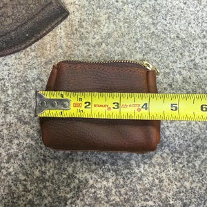 May include: A small, brown leather coin purse with a gold zipper, measuring approximately 4 inches in width. The purse is rectangular with rounded corners and a textured surface. A yellow tape measure is placed across the purse.