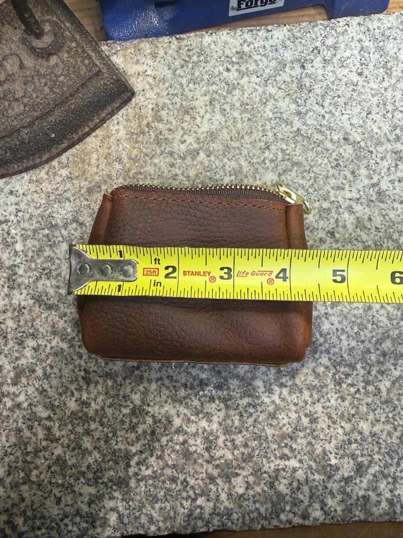 May include: A small, brown leather coin purse with a gold zipper, measuring approximately 4 inches in width. The purse is rectangular with rounded corners and a textured surface. A yellow tape measure is placed across the purse.