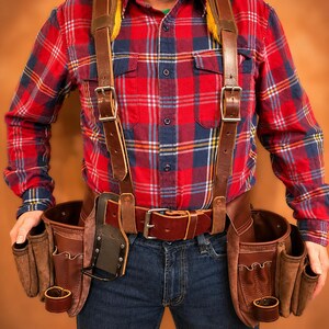 May include: A red, blue, and white plaid long-sleeve shirt worn with a brown leather tool belt and suspenders. The belt features multiple pouches, metal buckles, and a hammer loop. The suspenders have yellow padded shoulder straps and metal hardware.