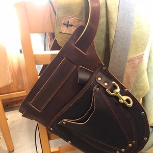 May include: A brown leather tool belt with a large pocket and a smaller pocket on the front. The belt has a brass clasp and is hanging on a wooden chair.