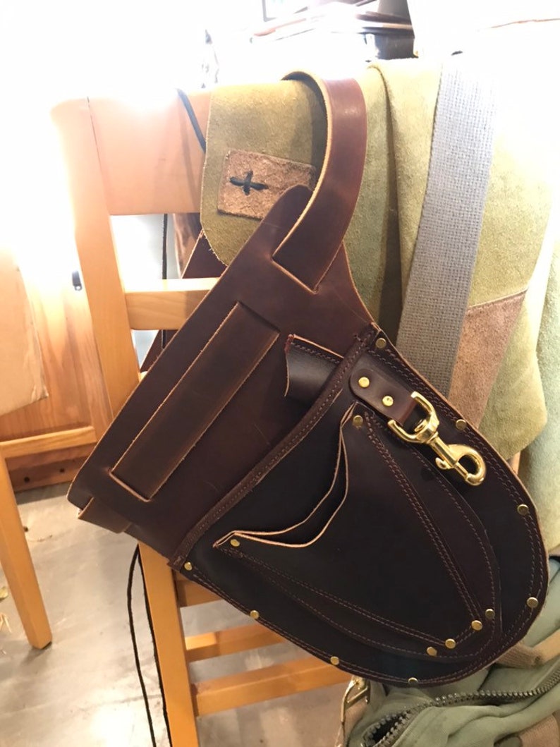 May include: A brown leather tool belt with a large pocket and a smaller pocket on the front. The belt has a brass clasp and is hanging on a wooden chair.