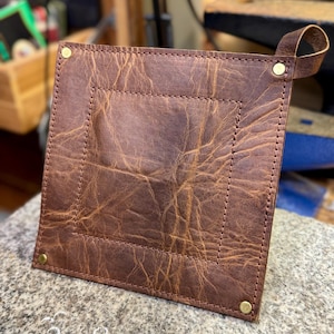 Puede incluir: Bandeja cuadrada de cuero marrón con un borde cosido y un asa de bucle. La bandeja está asegurada con remaches de latón en cada esquina. El cuero tiene un aspecto envejecido y texturizado.