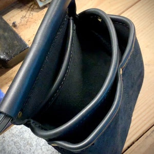 以下が含まれることがあります： 複数のポケットと真鍮のリベットが付いた黒い革製のツールバッグ。バッグは空で、滑らかで使い古した質感です。