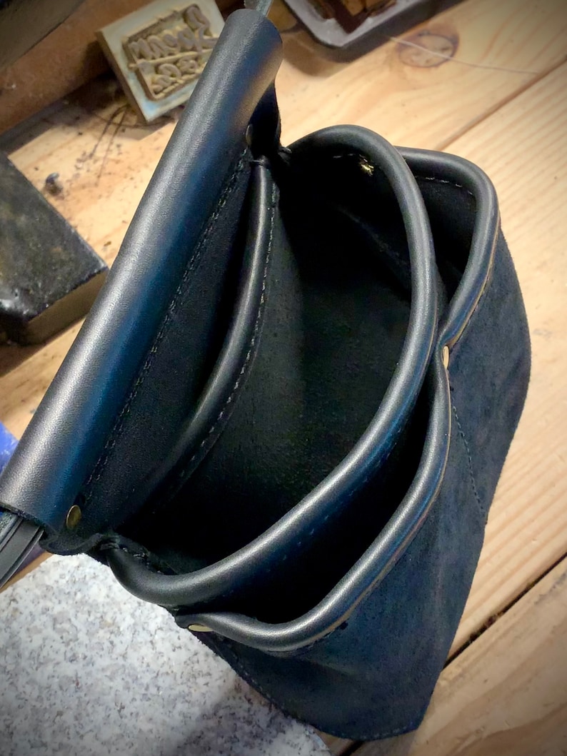 以下が含まれることがあります： 複数のポケットと真鍮のリベットが付いた黒い革製のツールバッグ。バッグは空で、滑らかで使い古した質感です。