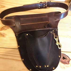 May include: A brown leather tool belt with a large pocket and a brass clip. The belt has a buckle and is secured with rivets.