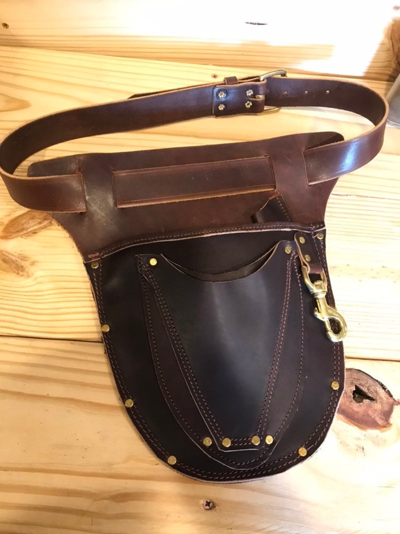 May include: A brown leather tool belt with a large pocket and a brass clip. The belt has a buckle and is secured with rivets.