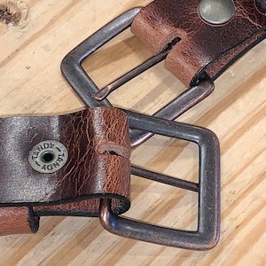 Peut inclure: Gros plan sur une ceinture en cuir marron avec deux boucles en laiton antique. La ceinture porte le logo Tandy sur la sangle.