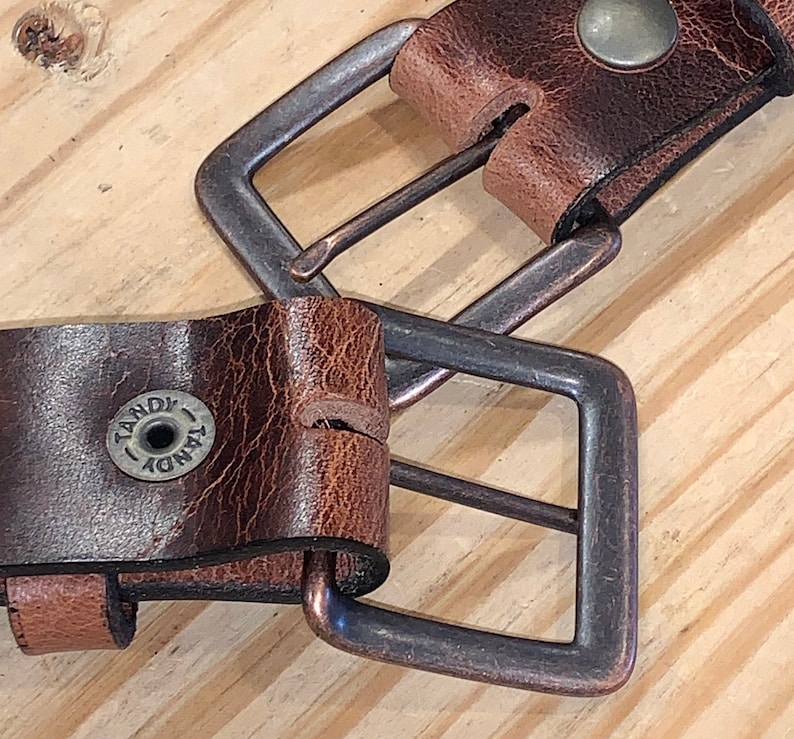 Peut inclure: Gros plan sur une ceinture en cuir marron avec deux boucles en laiton antique. La ceinture porte le logo Tandy sur la sangle.
