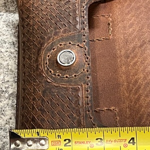 May include: Close-up of a brown leather phone case with a snap closure. The case is 2 inches wide. The case has a decorative embossed pattern and stitching.