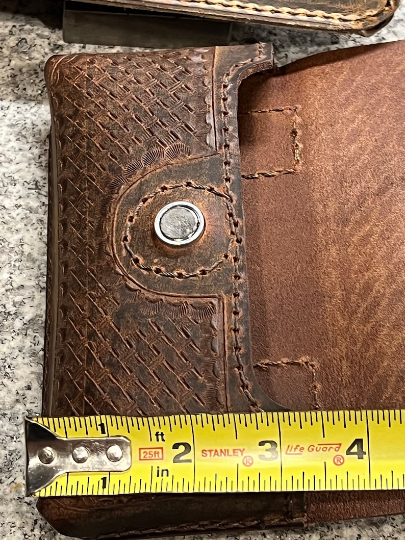 May include: Close-up of a brown leather phone case with a snap closure. The case is 2 inches wide. The case has a decorative embossed pattern and stitching.