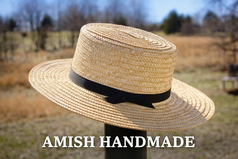 May include: A straw hat with a wide brim and a black band. The hat is handmade and features the text "AMISH HANDMADE" at the bottom. The hat is displayed outdoors.