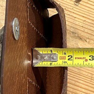 May include: A brown leather pouch with a flap and a snap closure. The pouch is being measured with a tape measure that reads 2 inches.