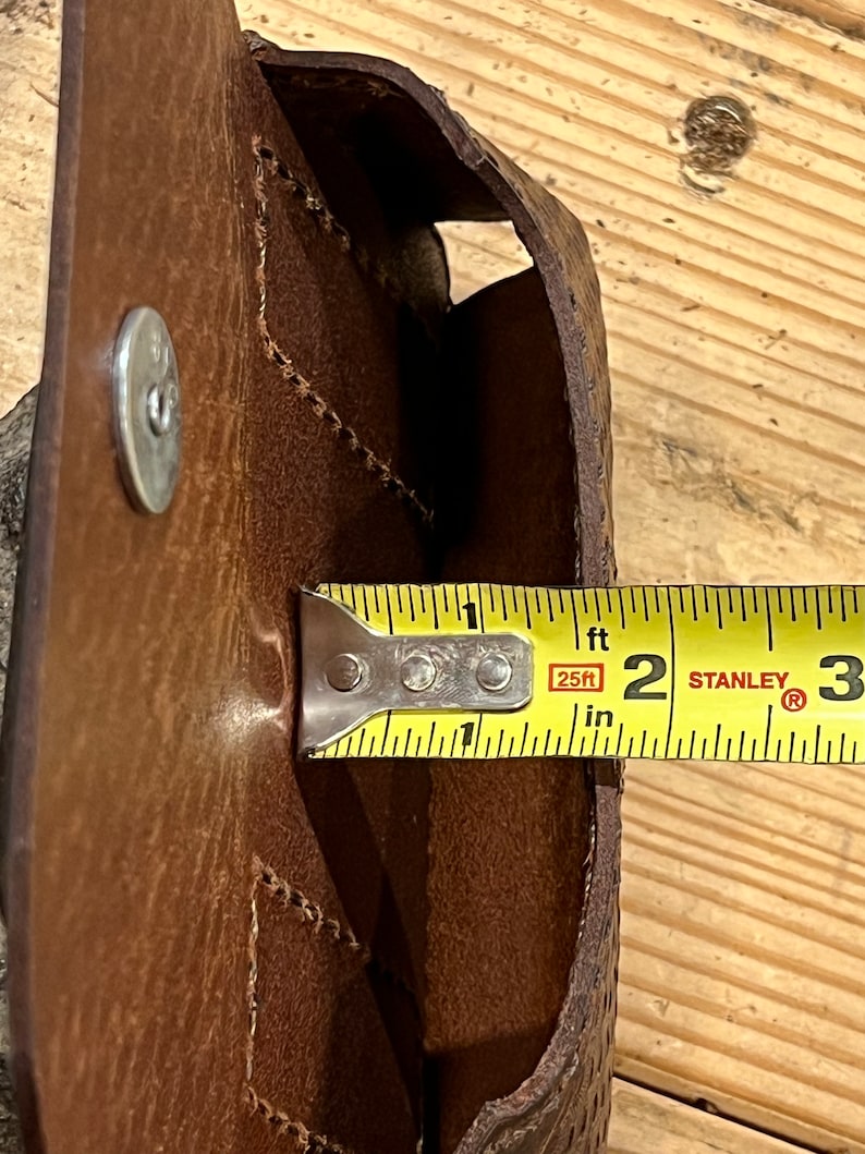 May include: A brown leather pouch with a flap and a snap closure. The pouch is being measured with a tape measure that reads 2 inches.