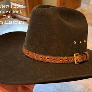 May include: A black cowboy hat with a brown leather band and gold-colored buckle. The hat has a wide brim and a rounded crown. The leather band is decorated with a floral pattern and secured with a gold-colored buckle.