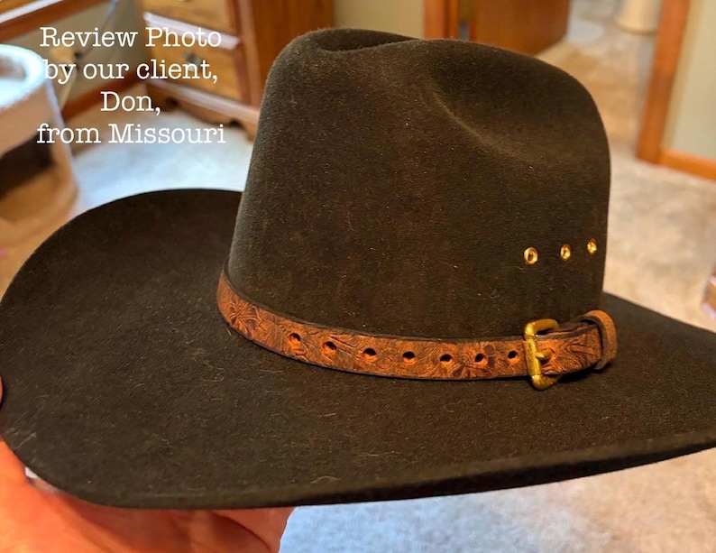 May include: A black cowboy hat with a brown leather band and gold-colored buckle. The hat has a wide brim and a rounded crown. The leather band is decorated with a floral pattern and secured with a gold-colored buckle.