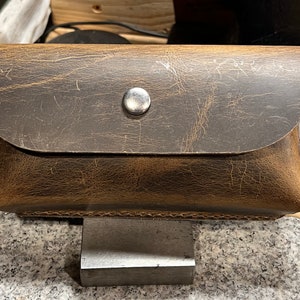 May include: A brown leather case with a metal button closure. The case has a distressed look and is made of high-quality leather.