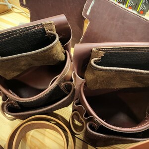 May include: Two brown leather tool belts with multiple pockets and compartments. The belts are empty and have a worn, vintage look.