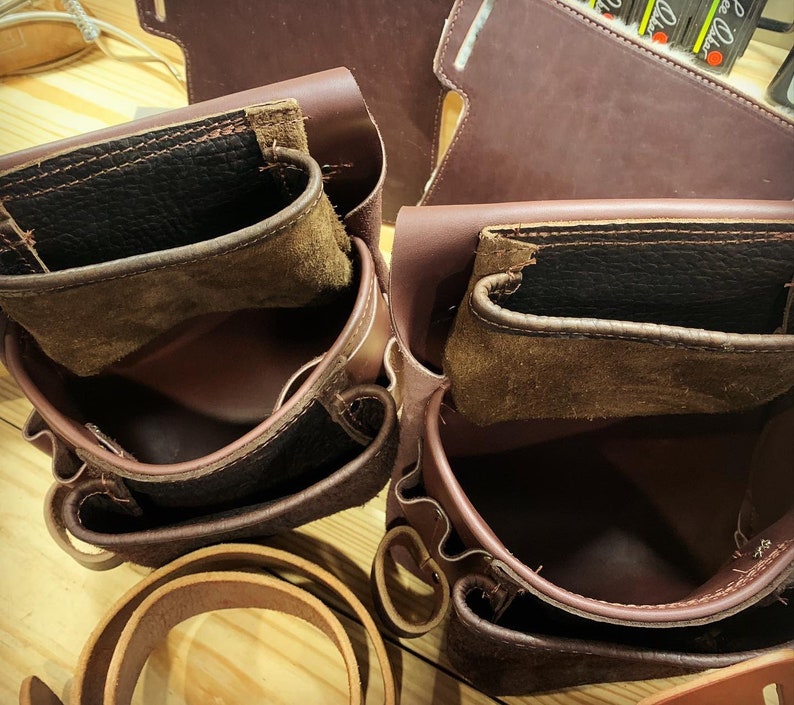May include: Two brown leather tool belts with multiple pockets and compartments. The belts are empty and have a worn, vintage look.