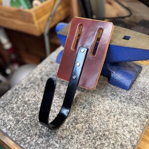 May include: A handmade leather and metal hook. The hook features a rectangular, reddish-brown leather backing with two vertical slots and a curved, black metal hook attached with three rivets. The hook is designed for hanging items.