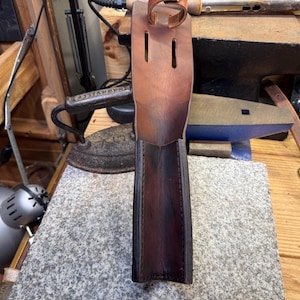 Puede incluir: Una funda de cuero hecha a mano, con un color marrón intenso y acentos más oscuros. La funda tiene un lazo y dos ranuras verticales en la parte superior. La imagen muestra la funda en un taller, con herramientas y otros artículos de cuero visibles.