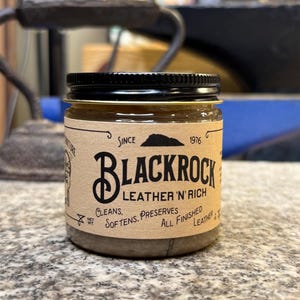 Puede incluir: Un frasco de vidrio con tapa negra y una etiqueta marrón. La etiqueta dice "Blackrock Leather 'n' Rich" y "Limpia, suaviza, conserva todo el cuero acabado".