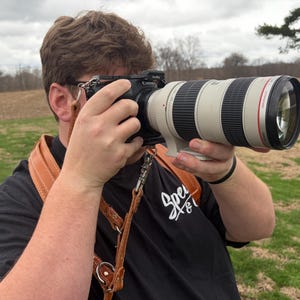 May include: A person holds a black camera with a large telephoto lens. The lens is white and black with red detailing. A brown leather camera strap is around the person's neck. The person wears a black t-shirt with white text.