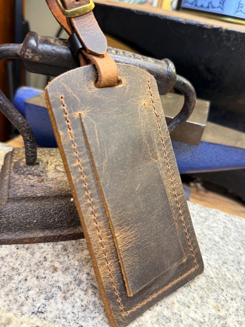 Op de afbeelding: Een bruine leren bagagelabel met een gestikte rand en een gespsluiting. De tag heeft een verweerde afwerking en een leren riem voor bevestiging. De tag is rechthoekig en heeft een vintage uitstraling.