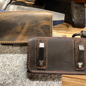 May include: Two leather phone cases, one brown and one dark brown, with stitching around the edges. The brown case has a distressed look and the dark brown case has two metal clips.