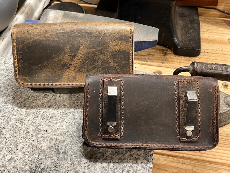 May include: Two leather phone cases, one brown and one dark brown, with stitching around the edges. The brown case has a distressed look and the dark brown case has two metal clips.