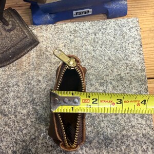 May include: A small, brown leather pouch with a gold zipper, measuring approximately 3 inches in length. The pouch is open, revealing a dark interior. A yellow measuring tape is placed across the opening.