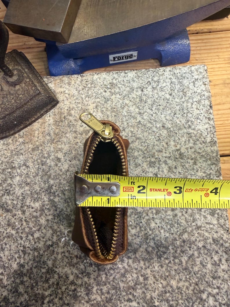 May include: A small, brown leather pouch with a gold zipper, measuring approximately 3 inches in length. The pouch is open, revealing a dark interior. A yellow measuring tape is placed across the opening.