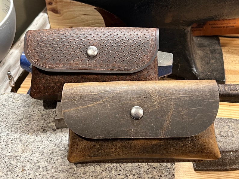May include: Two brown leather pouches with metal snaps. The top pouch has a textured design. The bottom pouch has a smooth surface.