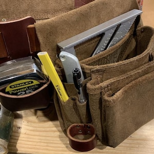 May include: A brown leather tool belt featuring multiple pouches and pockets. It contains a Stanley PowerLock 25' tape measure, a yellow Hanson pencil, a silver utility knife, a black pen, and a silver metal rafter square tucked inside.