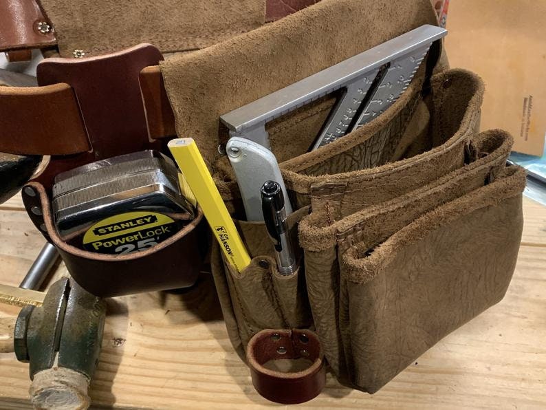 May include: A brown leather tool belt featuring multiple pouches and pockets. It contains a Stanley PowerLock 25' tape measure, a yellow Hanson pencil, a silver utility knife, a black pen, and a silver metal rafter square tucked inside.