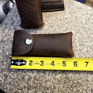 K&ouml;nnte beinhalten: Eine braune Lederscheide mit Druckknopfverschluss, etwa 15 cm lang. Die Scheide wird mit einem Ma&szlig;band gezeigt, und eine weitere &auml;hnliche Scheide ist im Hintergrund sichtbar. Das Leder hat ein abgenutztes Aussehen.