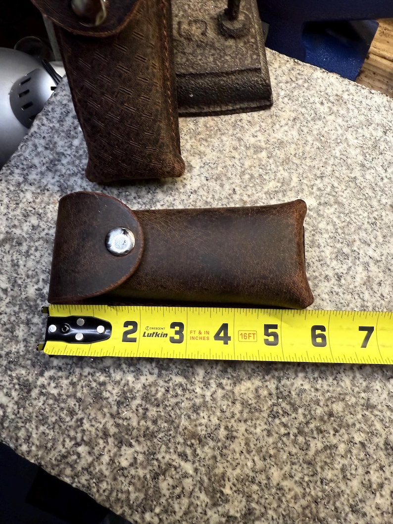 K&ouml;nnte beinhalten: Eine braune Lederscheide mit Druckknopfverschluss, etwa 15 cm lang. Die Scheide wird mit einem Ma&szlig;band gezeigt, und eine weitere &auml;hnliche Scheide ist im Hintergrund sichtbar. Das Leder hat ein abgenutztes Aussehen.