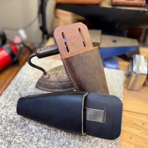 May include: A brown leather knife sheath with two slots and a black leather case with a metal clip. The items are on a speckled gray surface, with a vintage iron and other tools in the background.