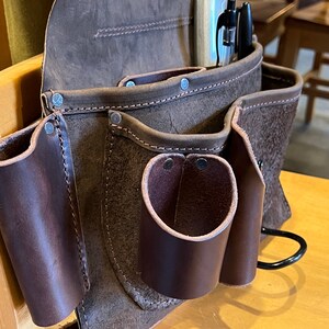 May include: A brown leather tool belt with multiple pockets and a metal hook. The belt has a worn, distressed look and is secured with rivets.