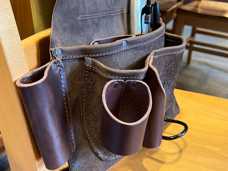 May include: A brown leather tool belt with multiple pockets and a metal hook. The belt has a worn, distressed look and is secured with rivets.
