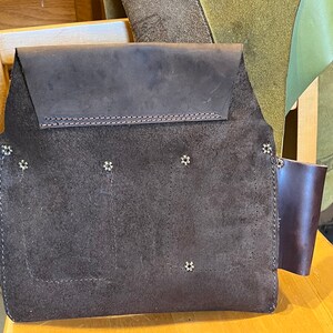 May include: A brown leather tool belt with multiple pockets and metal rivets. The belt has a suede-like texture and a brown leather strap on the right side.