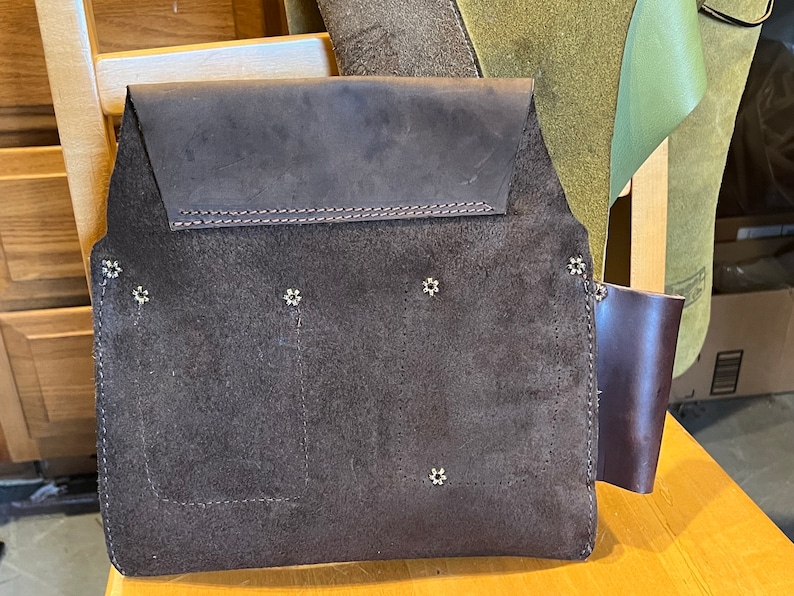 May include: A brown leather tool belt with multiple pockets and metal rivets. The belt has a suede-like texture and a brown leather strap on the right side.