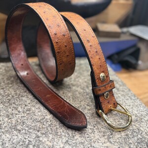 May include: A tan belt with a textured ostrich-like pattern and a dark brown smooth leather interior. The belt features a gold-toned metal buckle and two gold-toned metal rivets securing the loop near the buckle, with visible stitching along the edges.