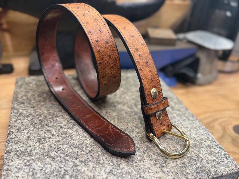May include: A tan belt with a textured ostrich-like pattern and a dark brown smooth leather interior. The belt features a gold-toned metal buckle and two gold-toned metal rivets securing the loop near the buckle, with visible stitching along the edges.