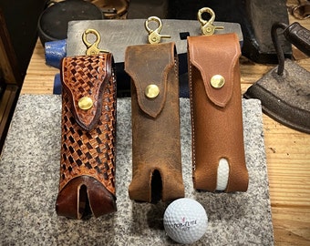 May include: Three leather golf ball holders with brass clasps and snaps. The holders are in different styles, one with a woven pattern, one with a distressed look, and one with a smooth finish. A white golf ball is in the foreground.