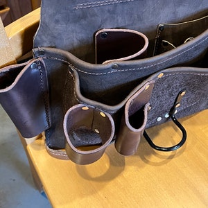 May include: A brown leather tool belt with multiple pockets and a metal loop. The belt has a worn, distressed look.