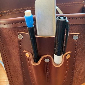 May include: A brown leather tool belt with two pockets. The pockets contain a silver utility knife, a black marker, and a white Sharpie marker.
