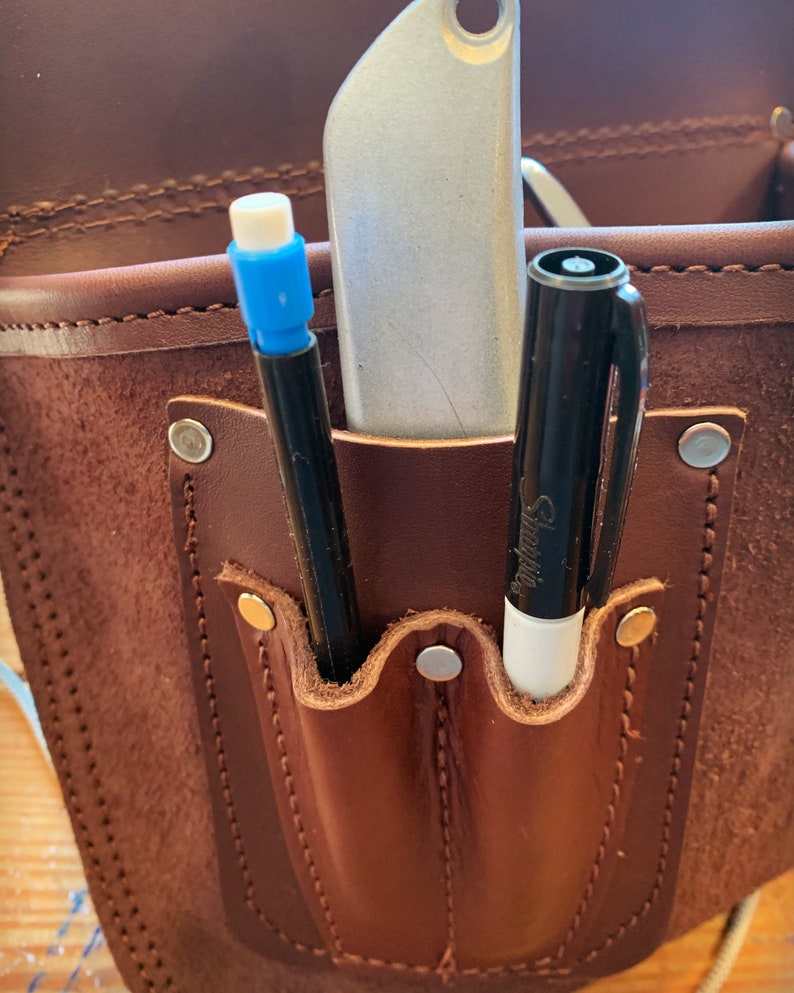 May include: A brown leather tool belt with two pockets. The pockets contain a silver utility knife, a black marker, and a white Sharpie marker.
