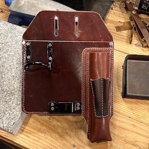 Op de afbeelding: Een bruine leren gereedschapsriem met een zak voor een gereedschap, een lus om gereedschap aan te hangen en een metalen gesp. De riem ligt op een houten oppervlak.